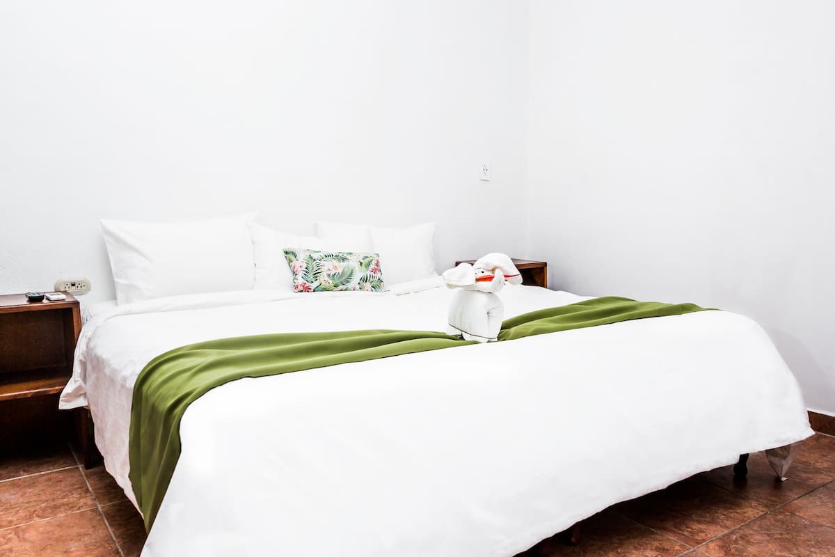 A neatly made bed with white bedding and a green throw, a wooden nightstand with a remote control, and a floral pillow.