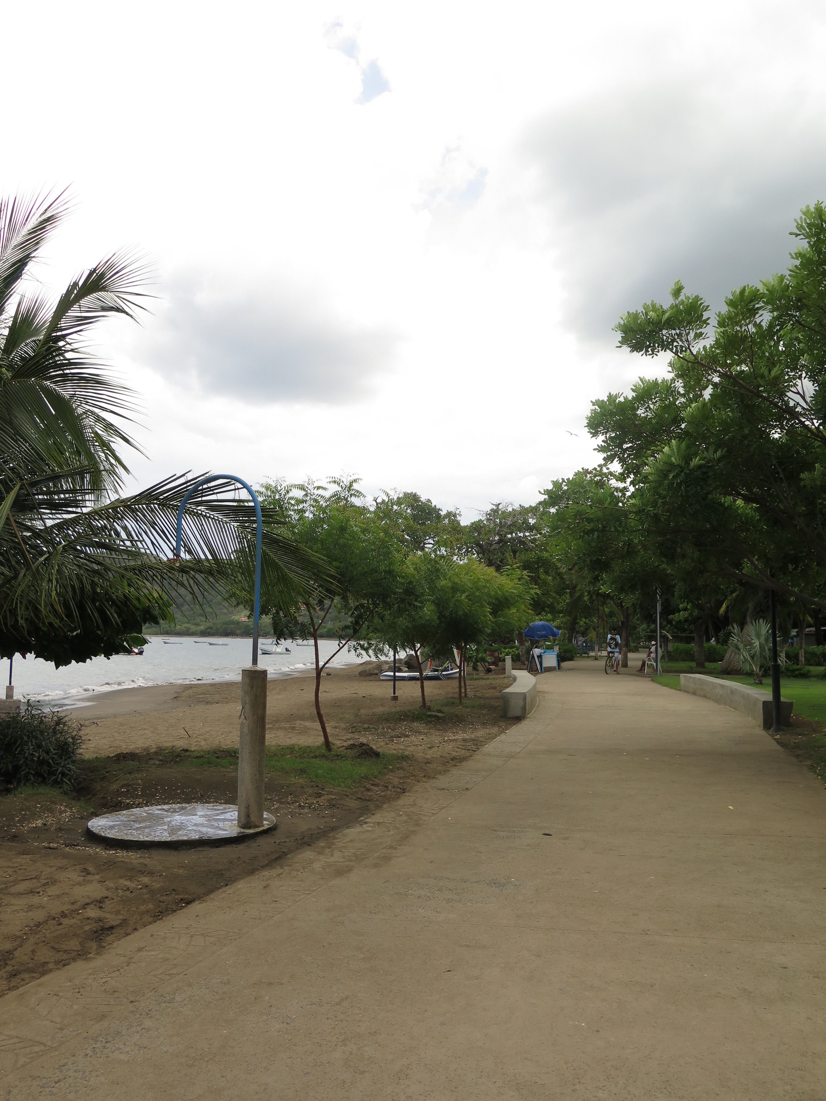 Un camino de hormigón que lleva a una playa con barcos en el agua, palmeras y un cielo nublado.