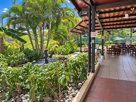 A covered outdoor seating area with tables and chairs, surrounded by lush greenery and a small water feature.