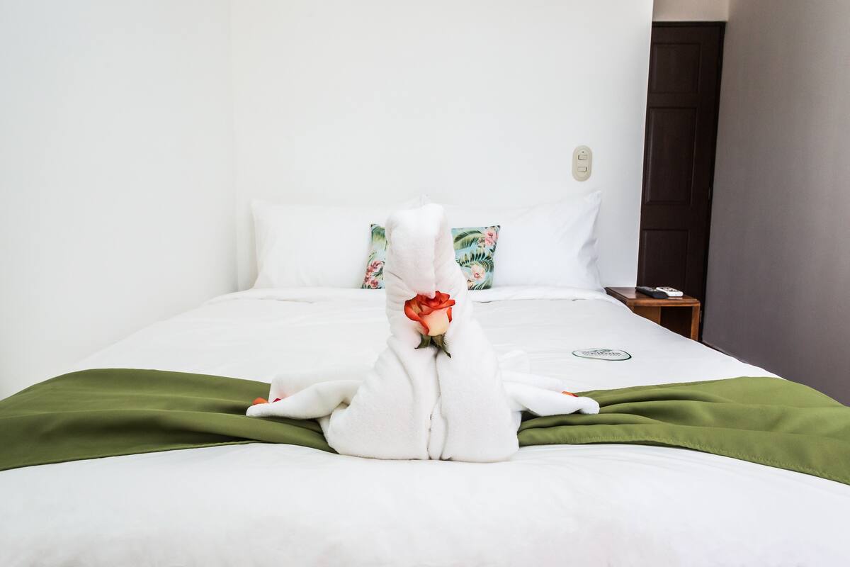 A neatly made bed with a green blanket, white pillows, and a towel sculpture holding a rose.