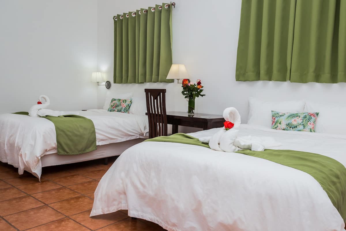 A hotel room with two beds, green curtains, a desk with a lamp, and a vase with flowers.