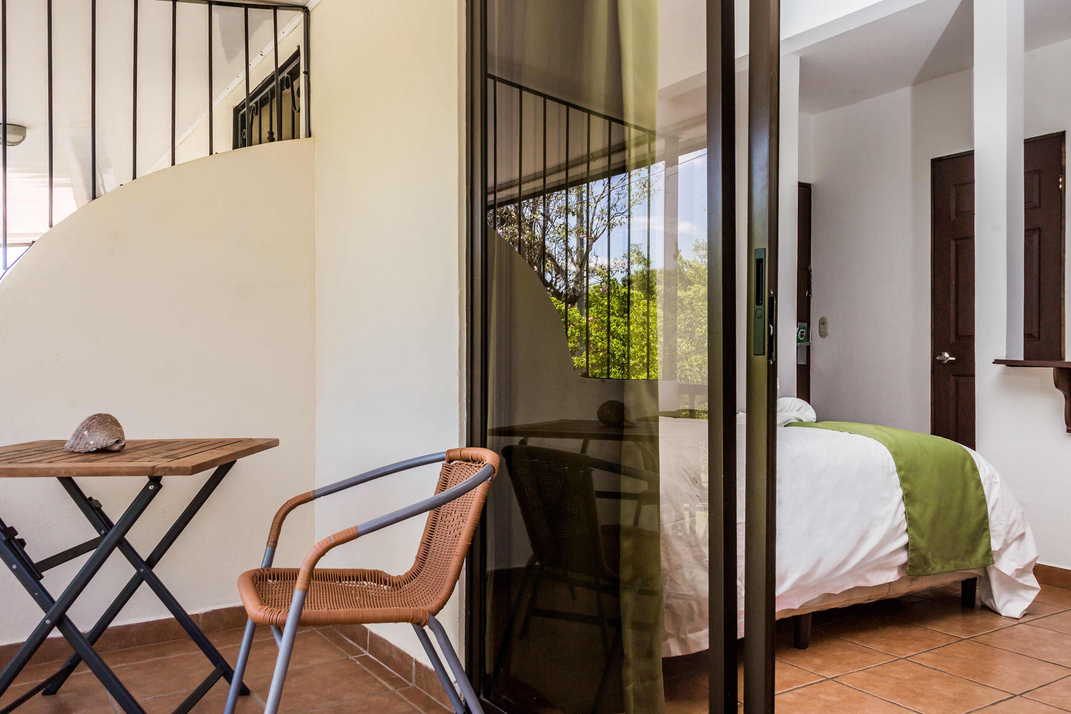 A balcony with a wicker chair, a small table, and a bed with a green blanket.
