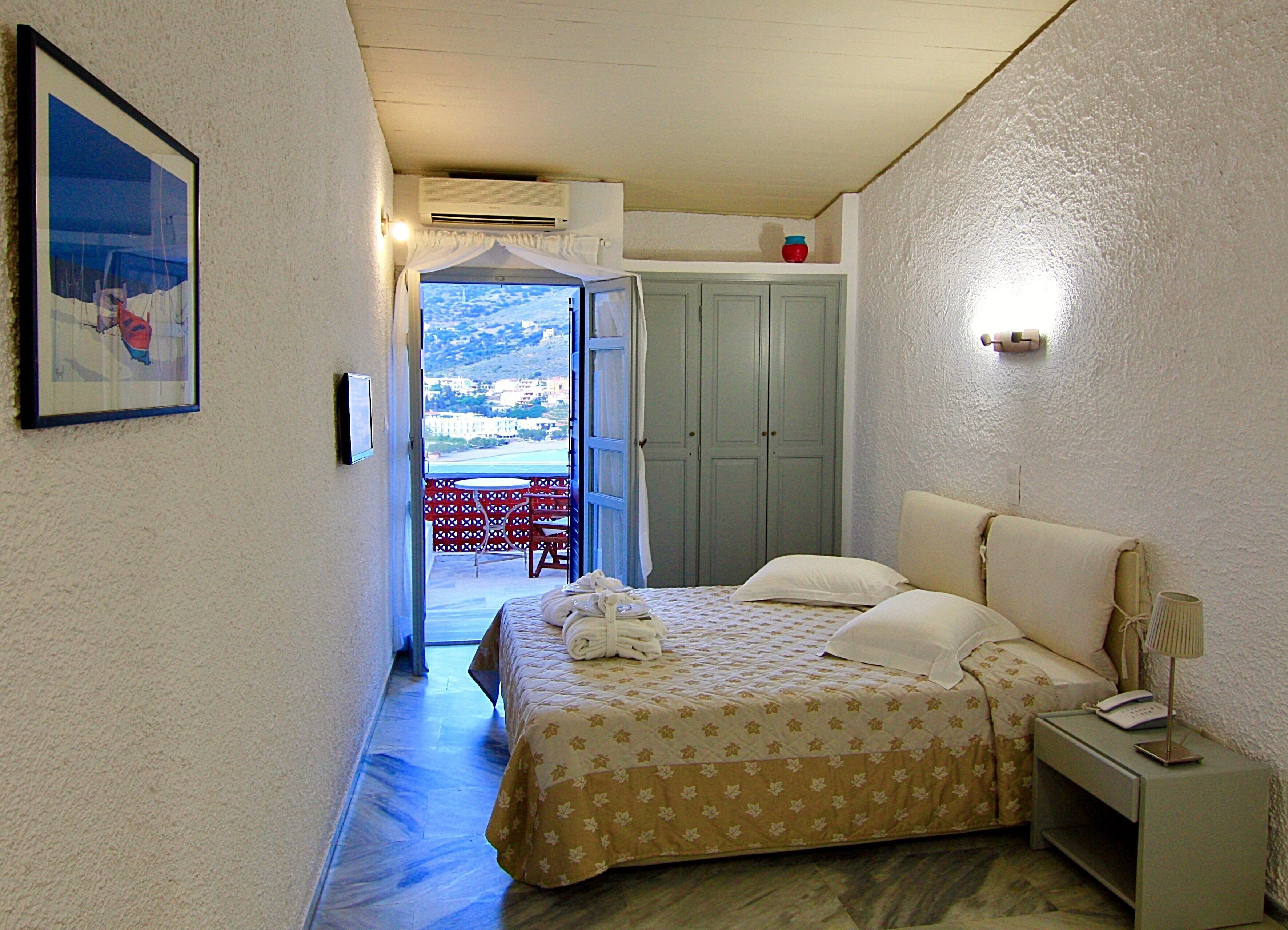 A bedroom with a bed, a nightstand, a wall-mounted lamp, a framed picture, a wardrobe, and an open door leading to a balcony.