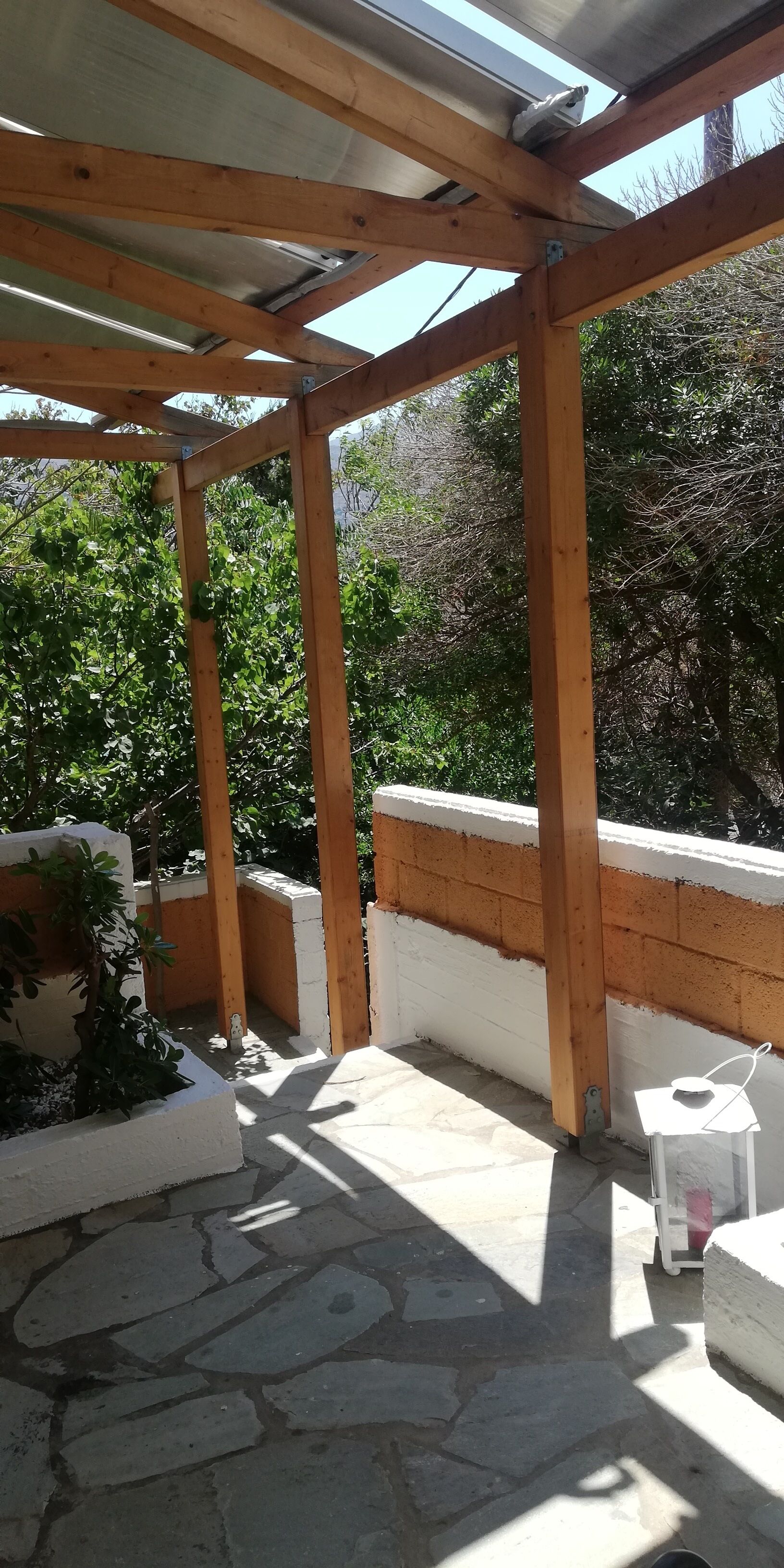 Un espacio al aire libre techado con estructura de madera, suelo de piedra y pared blanca.