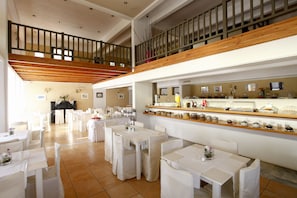Uma sala de jantar ampla, com várias mesas e cadeiras, uma escada de madeira e um balcão de buffet com diversos alimentos.