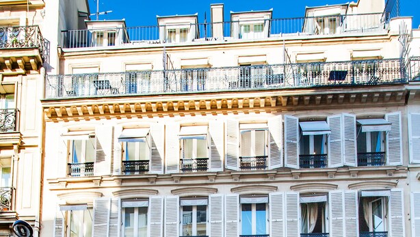 Un immeuble à plusieurs étages, doté de balcons et de devantures de magasins, avec un ciel bleu dégagé.