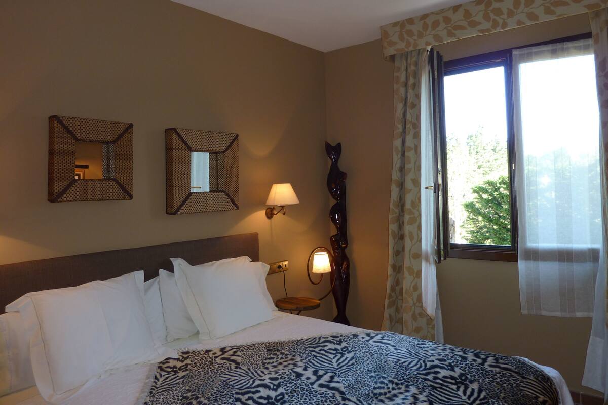 A hotel room with a bed, two wall-mounted mirrors, a nightstand with a lamp, and a window with curtains.