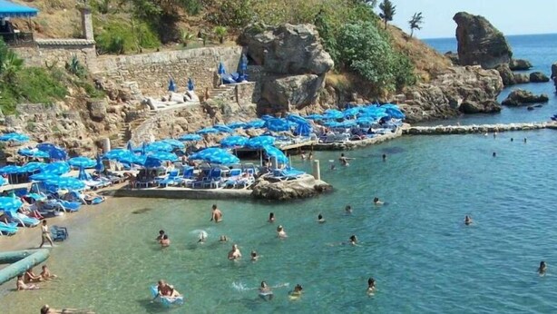 Una playa costera con agua cristalina, gente nadando y tomando el sol, y sombrillas azules.