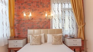 Un dormitorio con pared de ladrillo, una cama con ropa de cama blanca, dos mesitas de noche con lámparas y una ventana con cortinas.