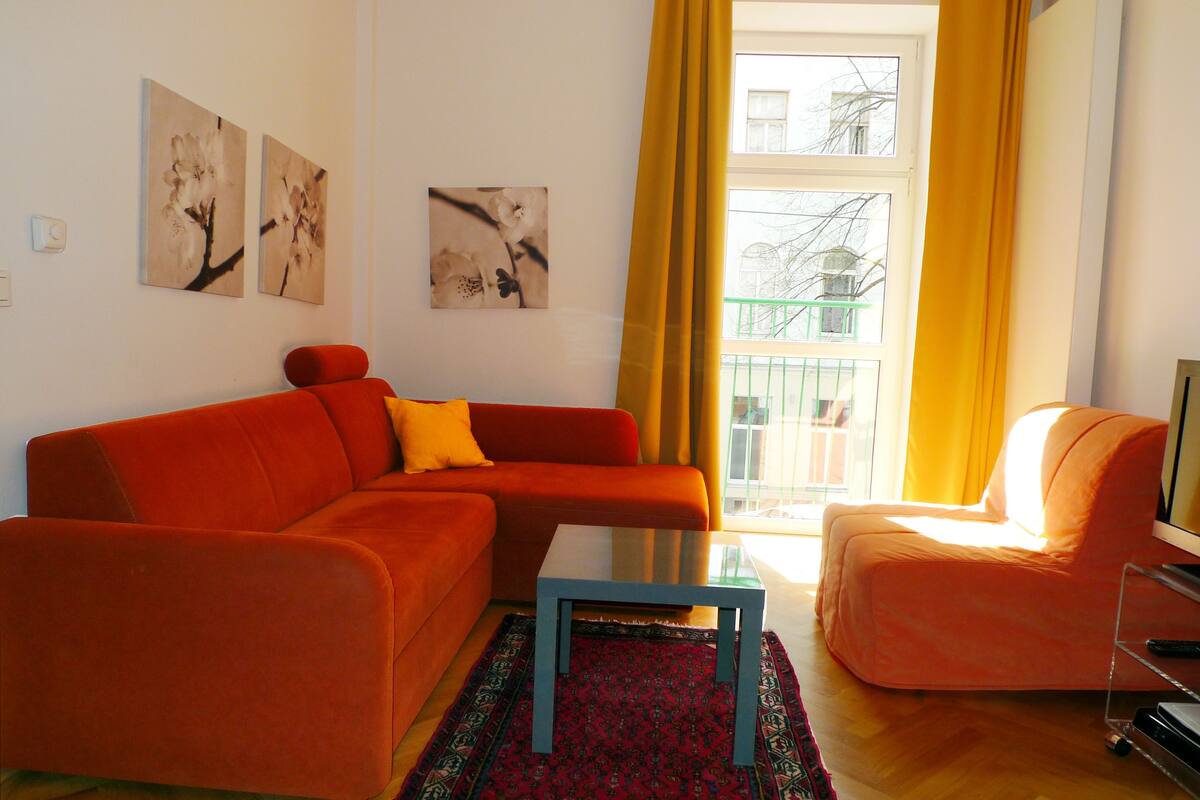 A living room with a sofa, a coffee table, and a chair. There are two wall-mounted paintings and a window with curtains.