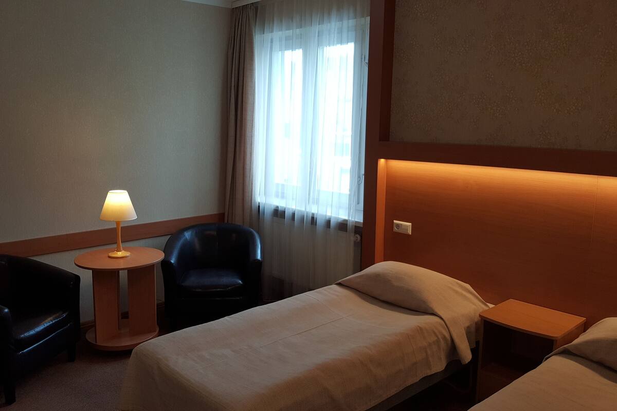 A hotel room with a bed, a chair, a small table with a lamp, and a window with curtains.