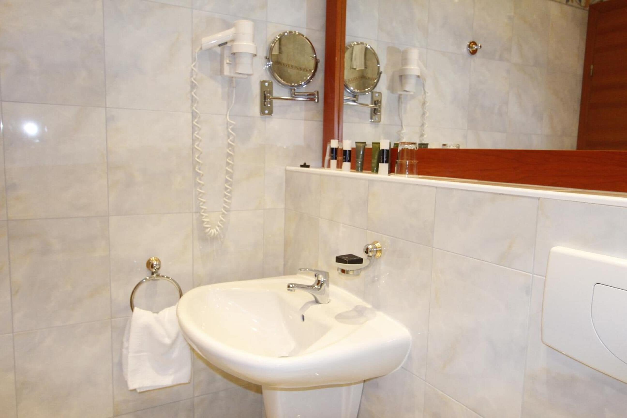 A bathroom with a white sink, two mirrors, a towel rack, and toiletries on a shelf.