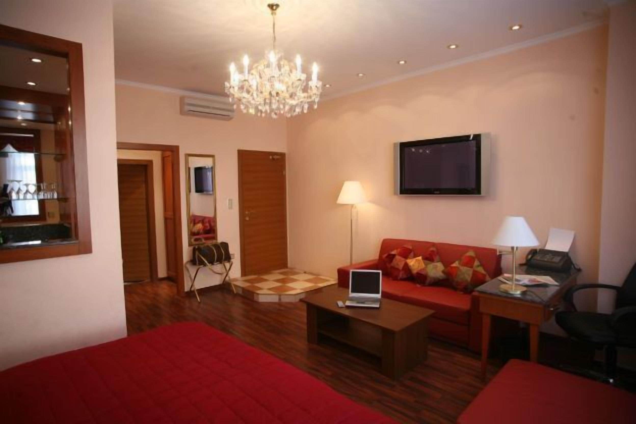 Une chambre d’hôtel avec un canapé rouge, un ordinateur portable posé sur une table basse, un téléviseur fixé au mur et un lustre.
