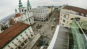 A historic square with traditional architecture, including a church with green spires and a central fountain. - Hotel Corvinus (Vienna)