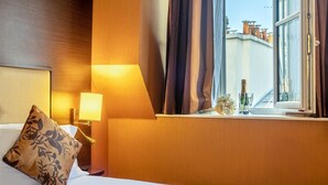 A hotel room with a bed, a nightstand, a lamp, a window with curtains, and a view of buildings outside. - Hotel Saint Honore 85 (Paris)