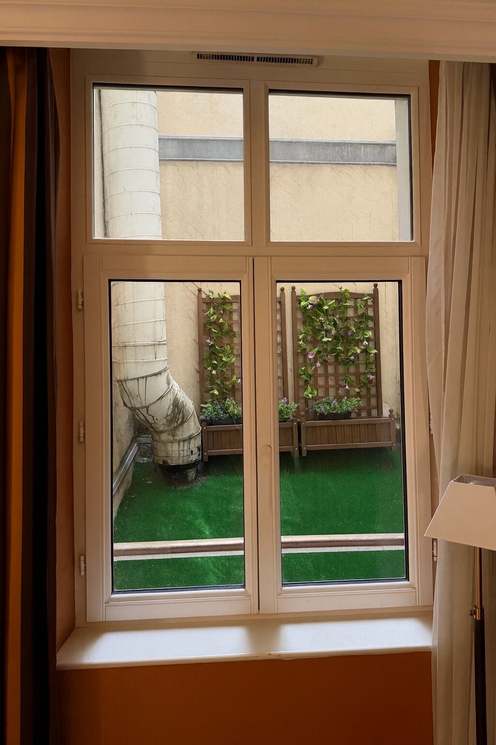 A window with a view of a courtyard with green artificial grass, wooden planters with flowers, and a white pillar.