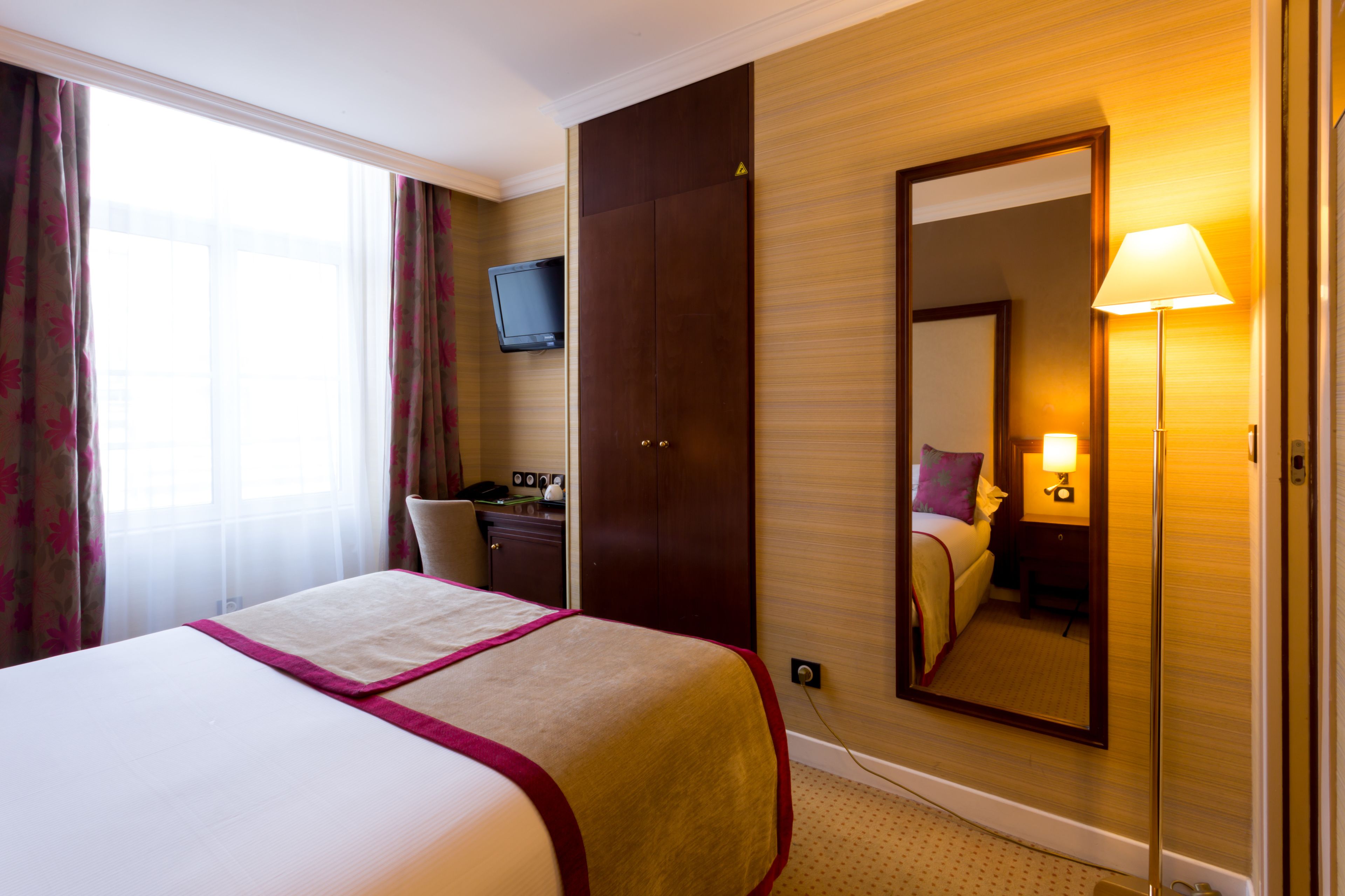 A hotel room with a bed, a desk, a television, a mirror, and a lamp.