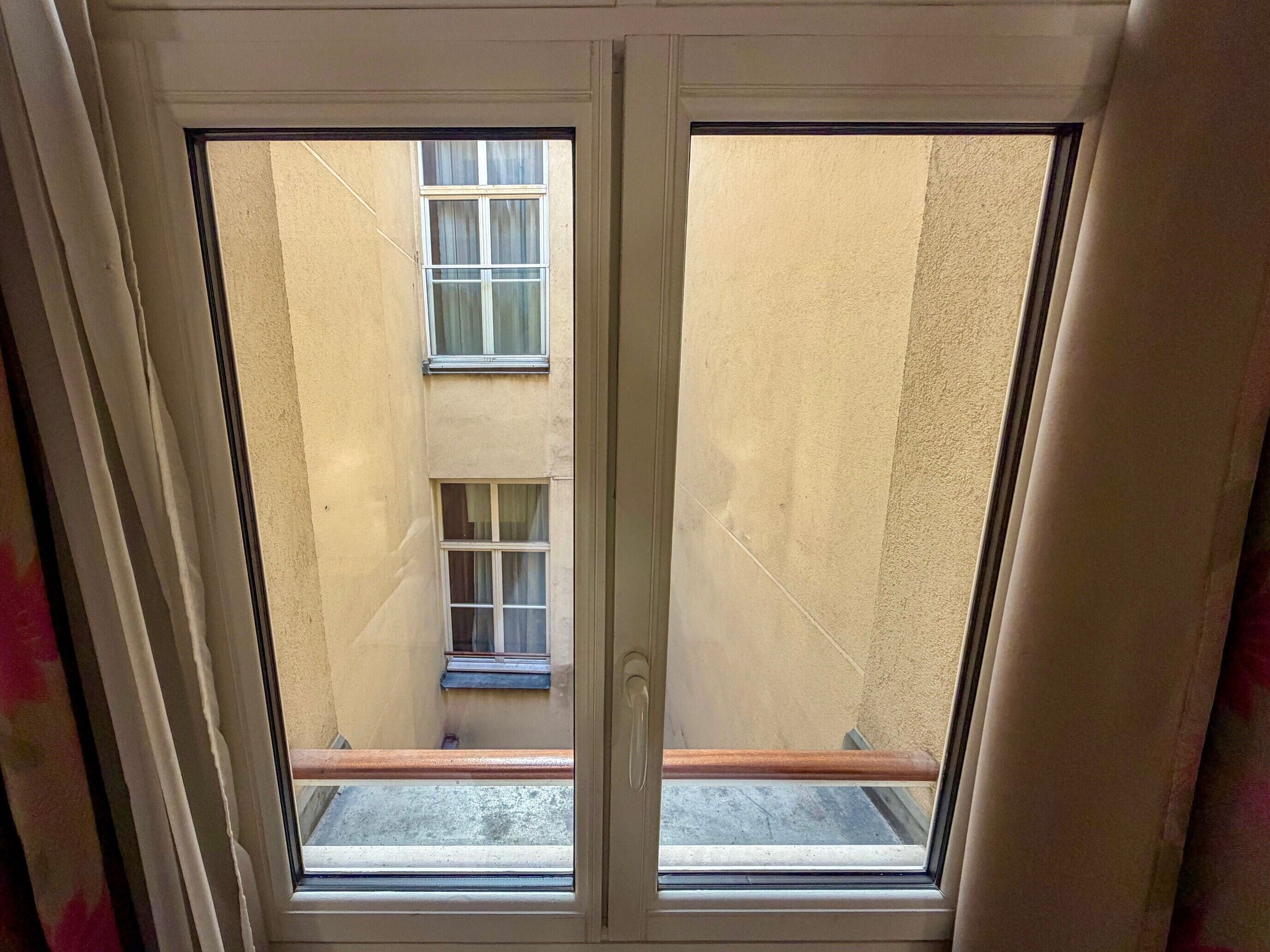 Une vue à travers une fenêtre d’un bâtiment, où l’on aperçoit deux autres fenêtres et un petit balcon.