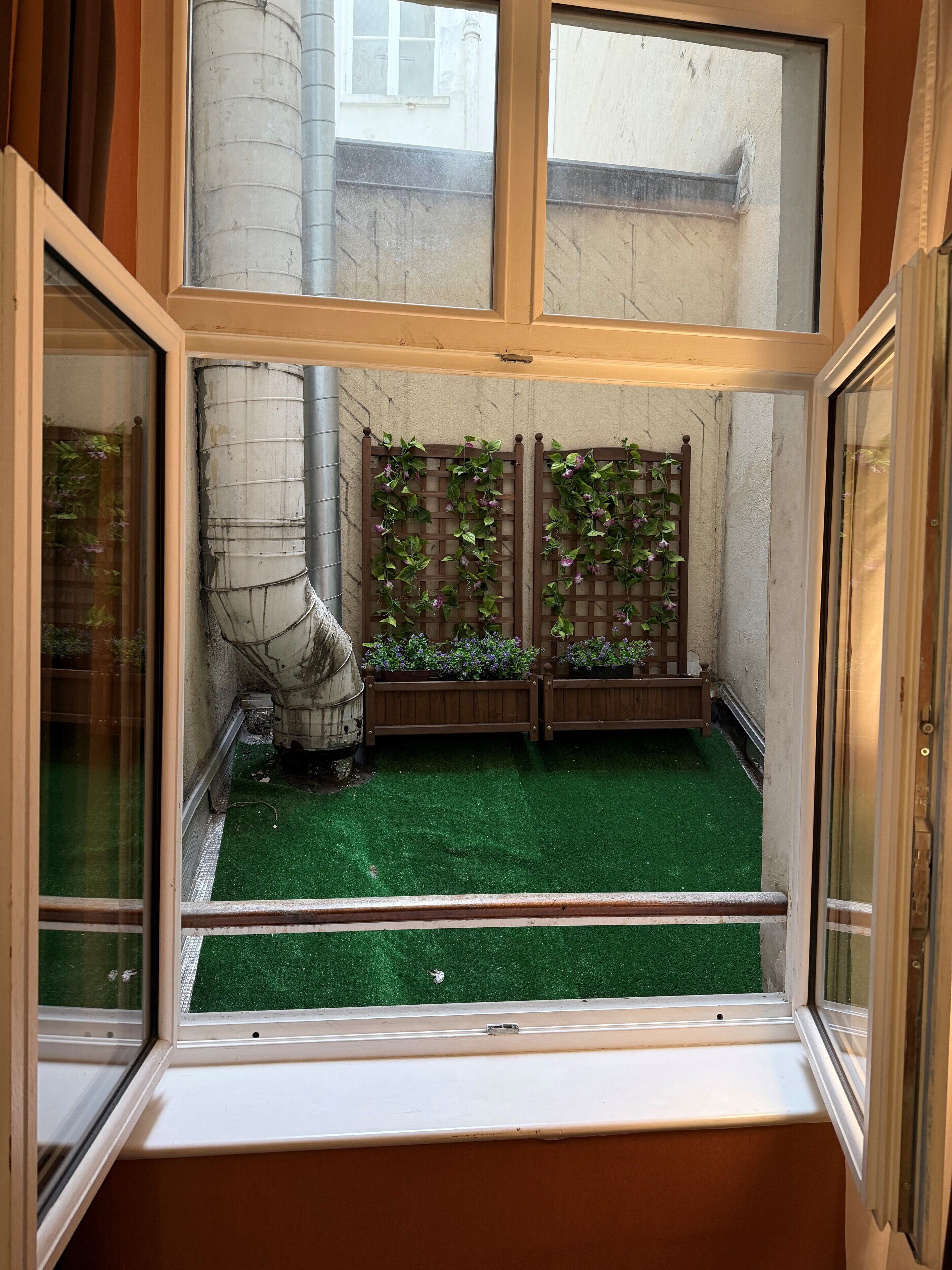 A view from a window into a room with a green artificial turf floor, a wooden lattice wall with climbing plants, and a metal pipe structure.