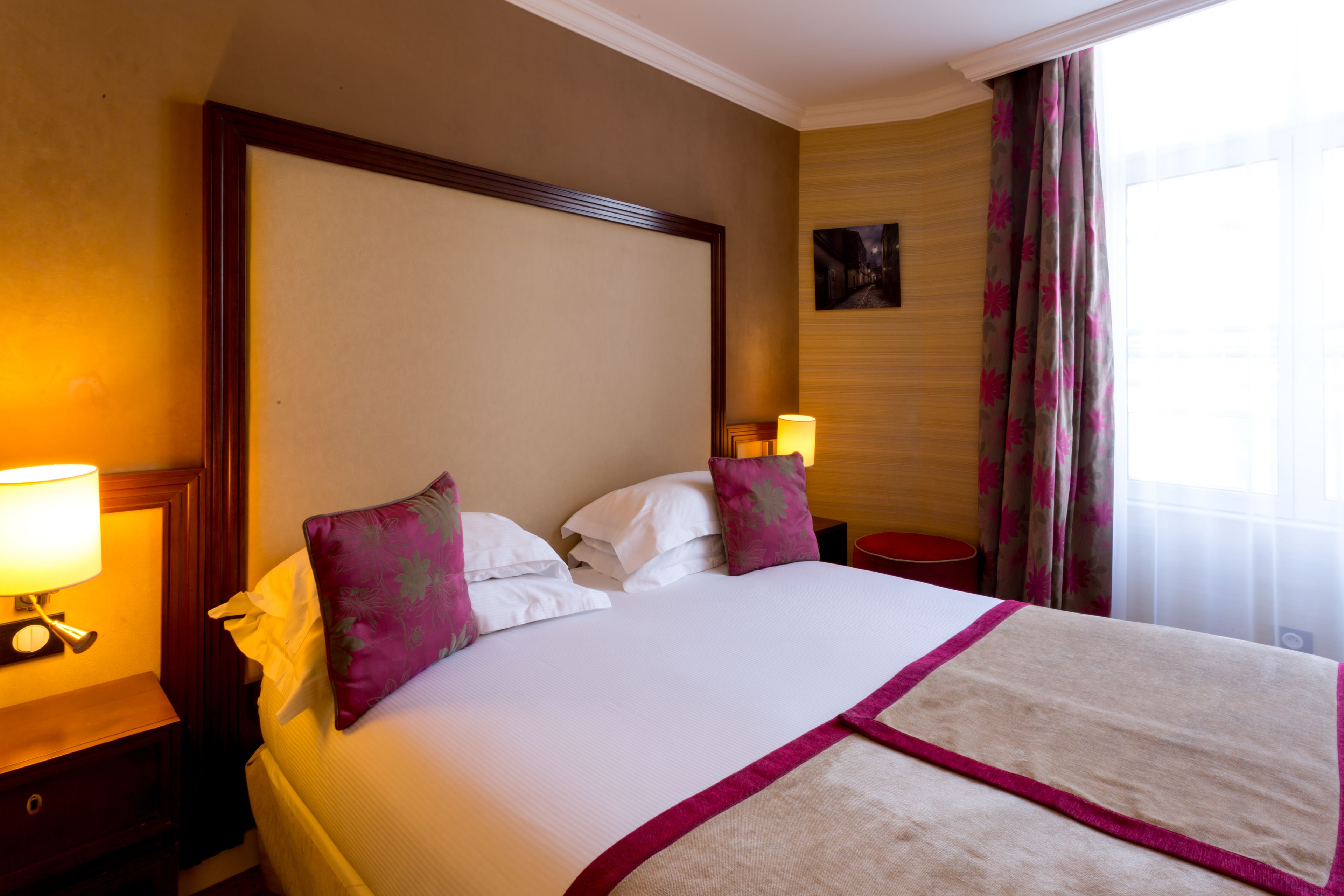 Un cuarto de hotel con una cama grande, dos mesitas de noche, una lámpara de noche, un cuadro en la pared y una ventana con cortinas.