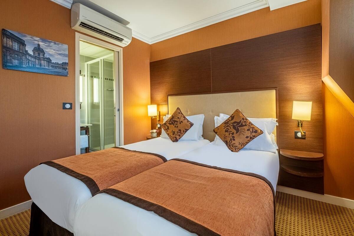 A hotel room with a large bed, two bedside tables with lamps, an air conditioning unit, and a painting on the wall.