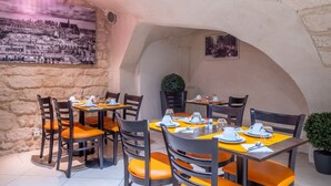 A restaurant with stone walls and a mural, tables set with orange cushions and plates. - Hotel Saint Honore 85 (Paris)