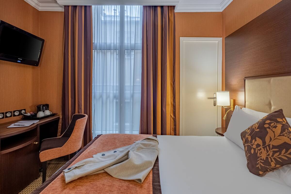 A hotel room with a bed, a desk with a chair, a television, and a window with curtains.
