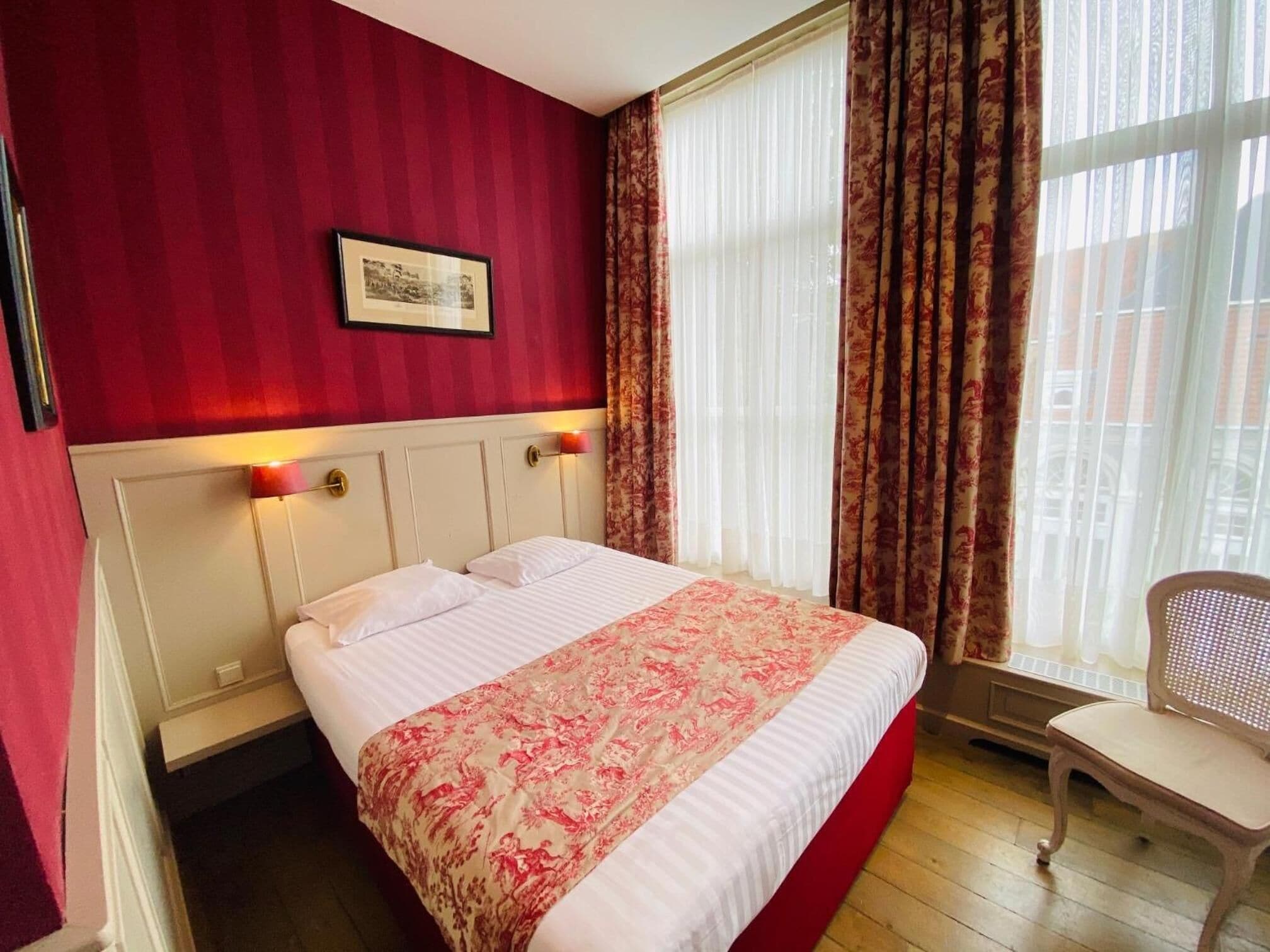 Une chambre d’hôtel avec un lit, une chaise, une fenêtre avec des rideaux et un tableau encadré au mur.