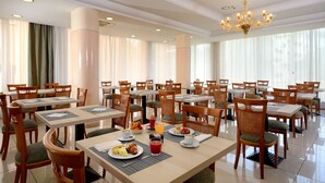 Comedor de hotel con varias mesas y sillas, servicio de desayuno y amplias ventanas con cortinas.
