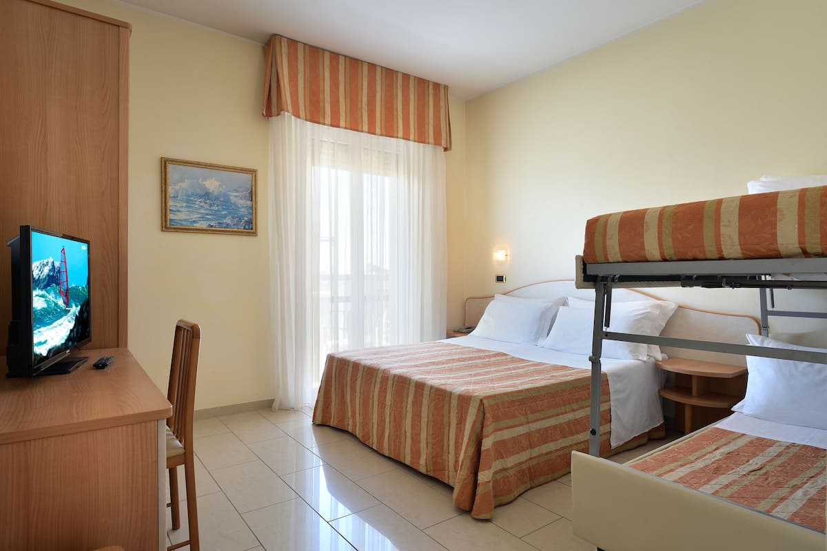 Une chambre d’hôtel avec un lit superposé, un bureau avec une télévision, une chaise, un lit avec une couvre-lit à rayures, une table de chevet et une fenêtre avec des rideaux.