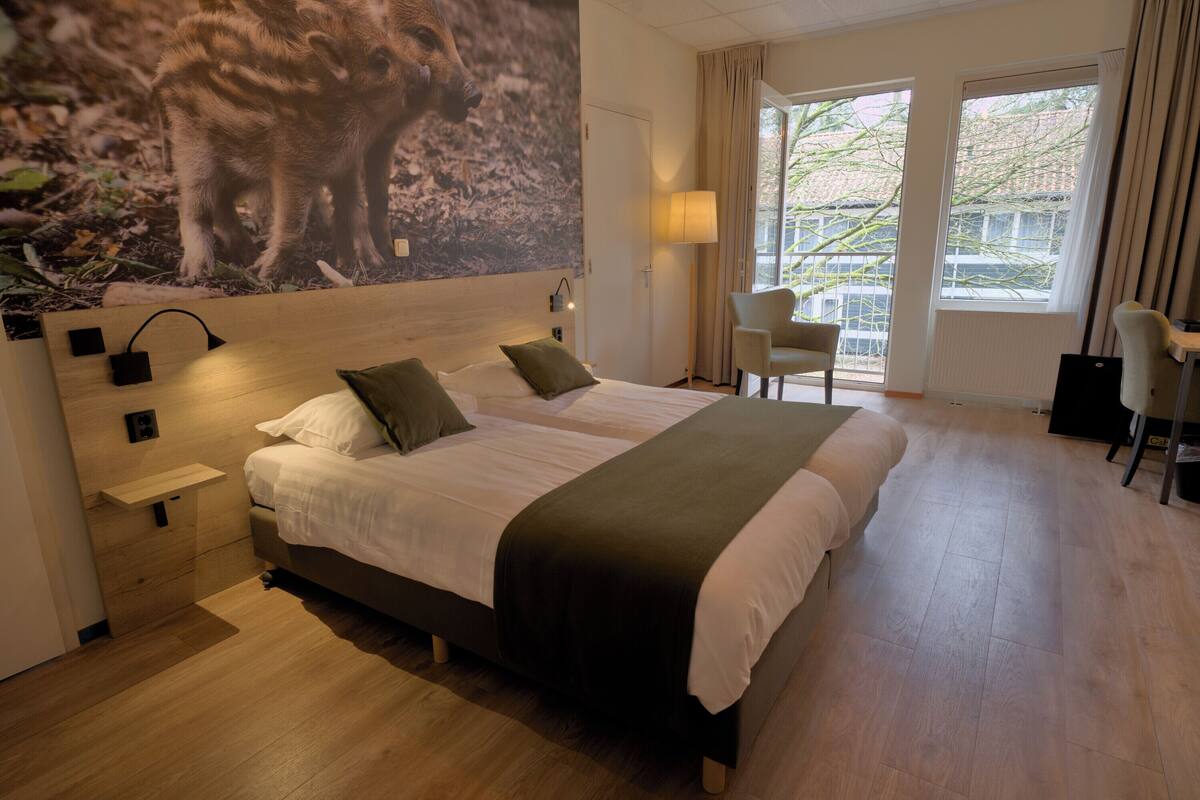 A hotel room with a large bed, a nightstand, a lamp, and a view of the outdoors through the window.