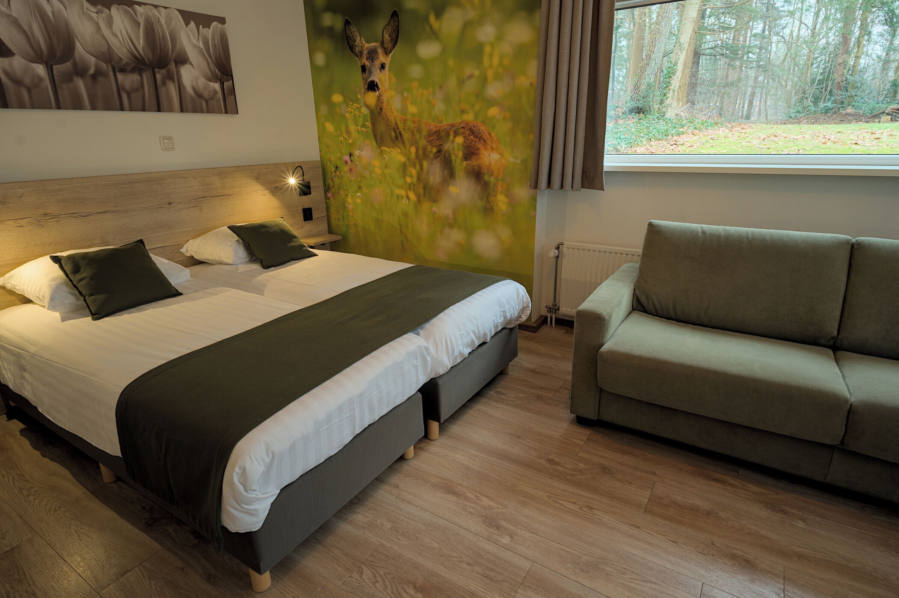 Ein Hotelzimmer mit einem großen Bett, einem grünen Sofa, einem Wandbild von einem Reh auf einer Wiese und einem Fenster mit Blick auf einen Wald.