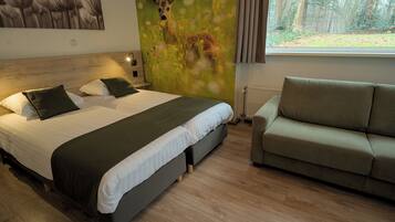 Ein Hotelzimmer mit einem großen Bett, einem grünen Sofa, einem Wandbild von einem Reh auf einer Wiese und einem Fenster mit Blick auf einen Wald.