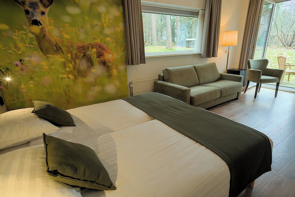 A hotel room with a large bed, a sofa, a chair, a window with a view of trees, and a wall mural of a deer.