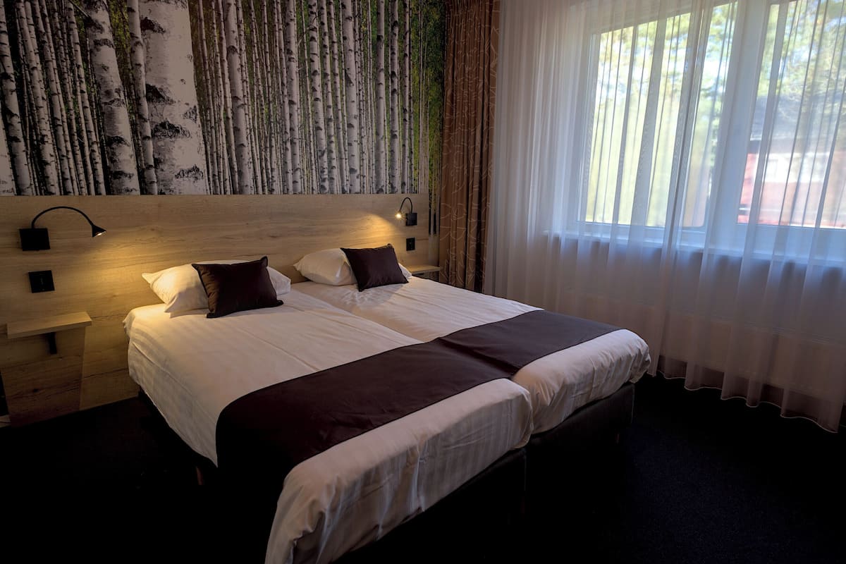 A hotel room with a large bed, two bedside tables, a wall-mounted lamp, and a view of trees through sheer curtains.