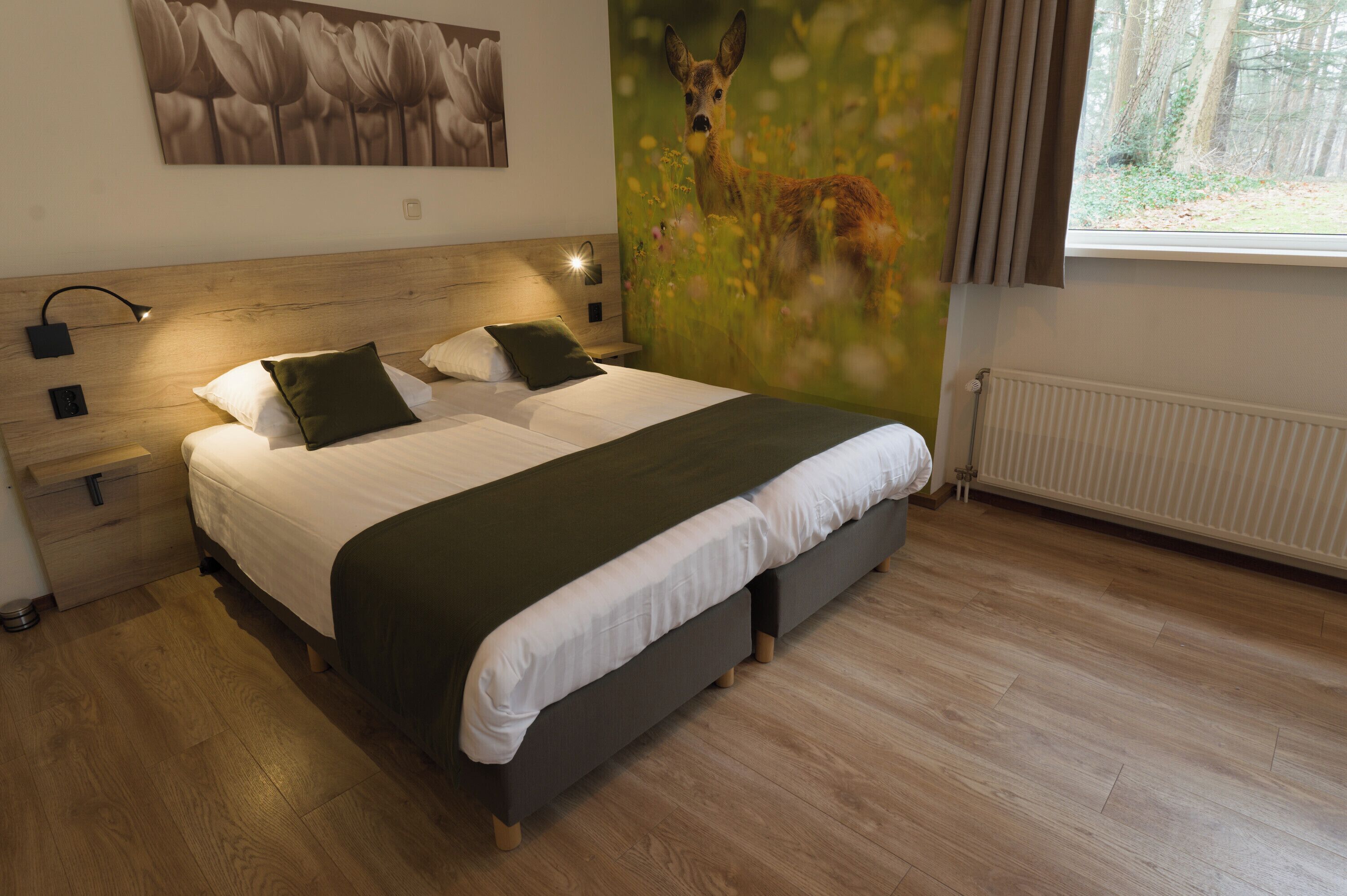 Un dormitorio moderno con cabecera de madera, una cama grande con ropa blanca y almohadas verdes, una mesita de noche con lámpara, y un mural en la pared con un ciervo en un campo.