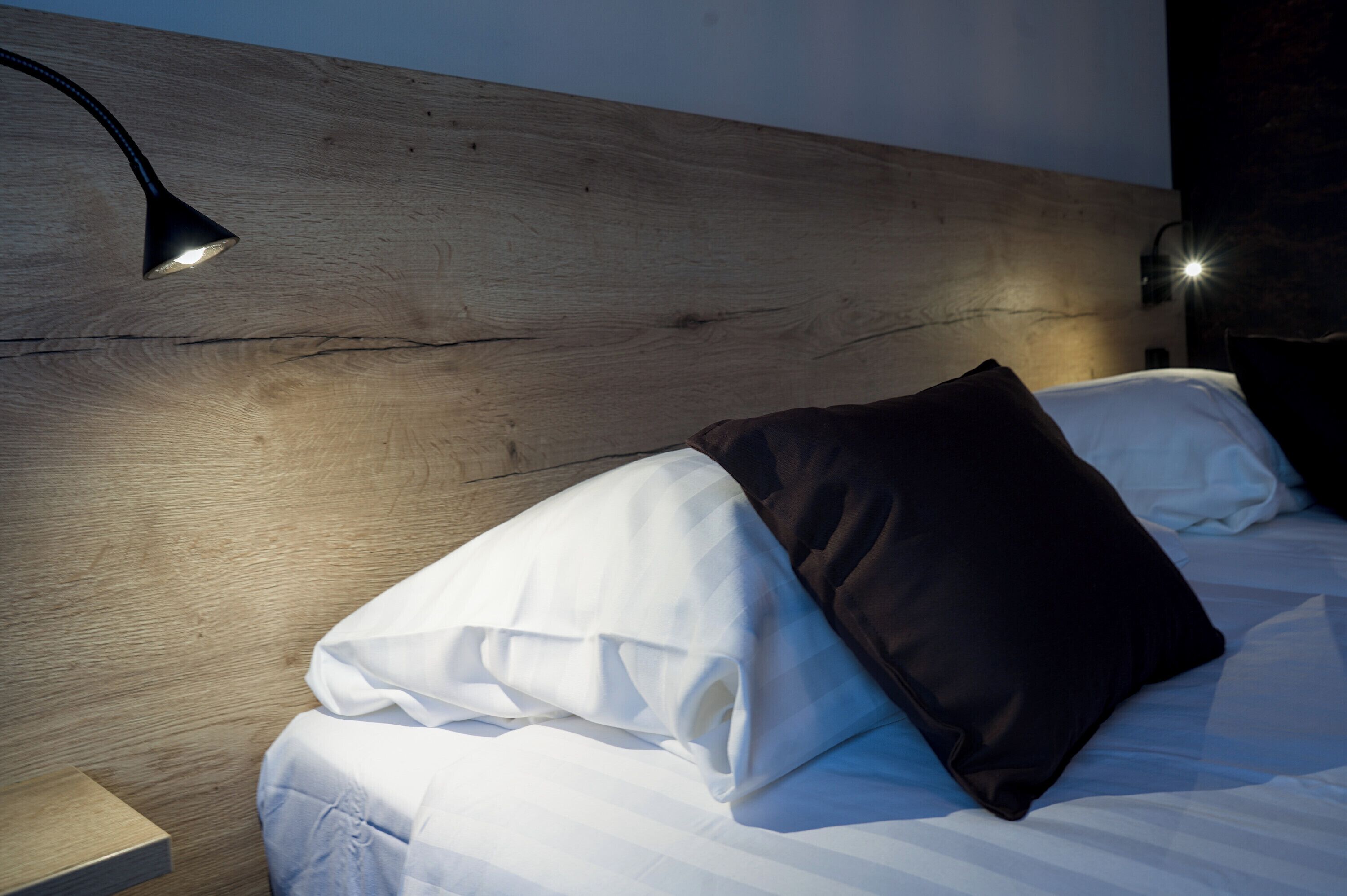 A neatly made bed with white and dark pillows, a wooden headboard, and a bedside lamp.