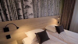 Une chambre d’hôtel avec une tête de lit en bois, deux lits avec une literie blanche et marron foncé, une table de chevet fixée au mur avec une lampe, et un mur recouvert d’un papier peint représentant un bouleau.