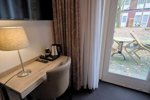 Habitación de hotel con escritorio, lámpara, control remoto, cafetera y vistas al exterior.