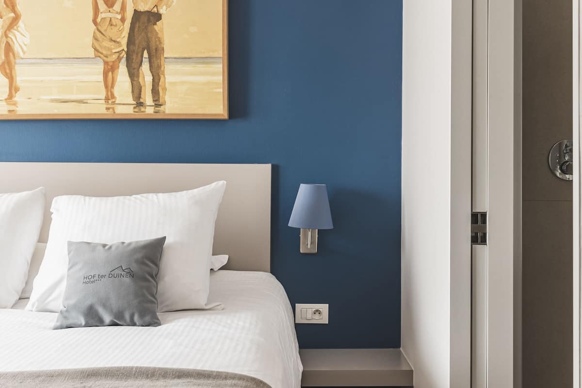 A hotel room with a bed, a painting of a couple on a beach, a nightstand with a lamp, and a door to a shower area.