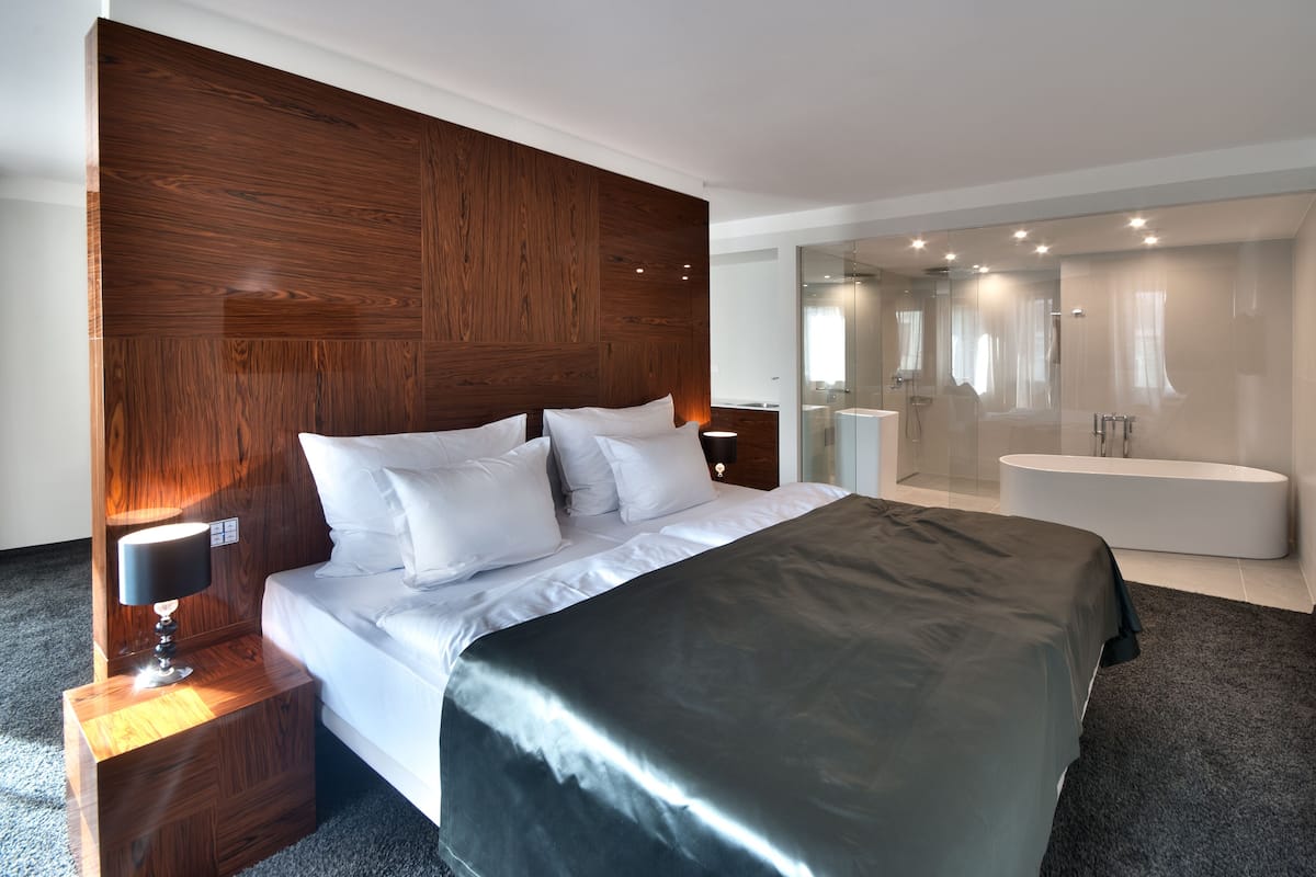 Une chambre d’hôtel moderne dotée d’un grand lit, d’une tête de lit en bois, d’une table de chevet avec une lampe et d’une salle de bain fermée par des parois de verre, avec une baignoire.