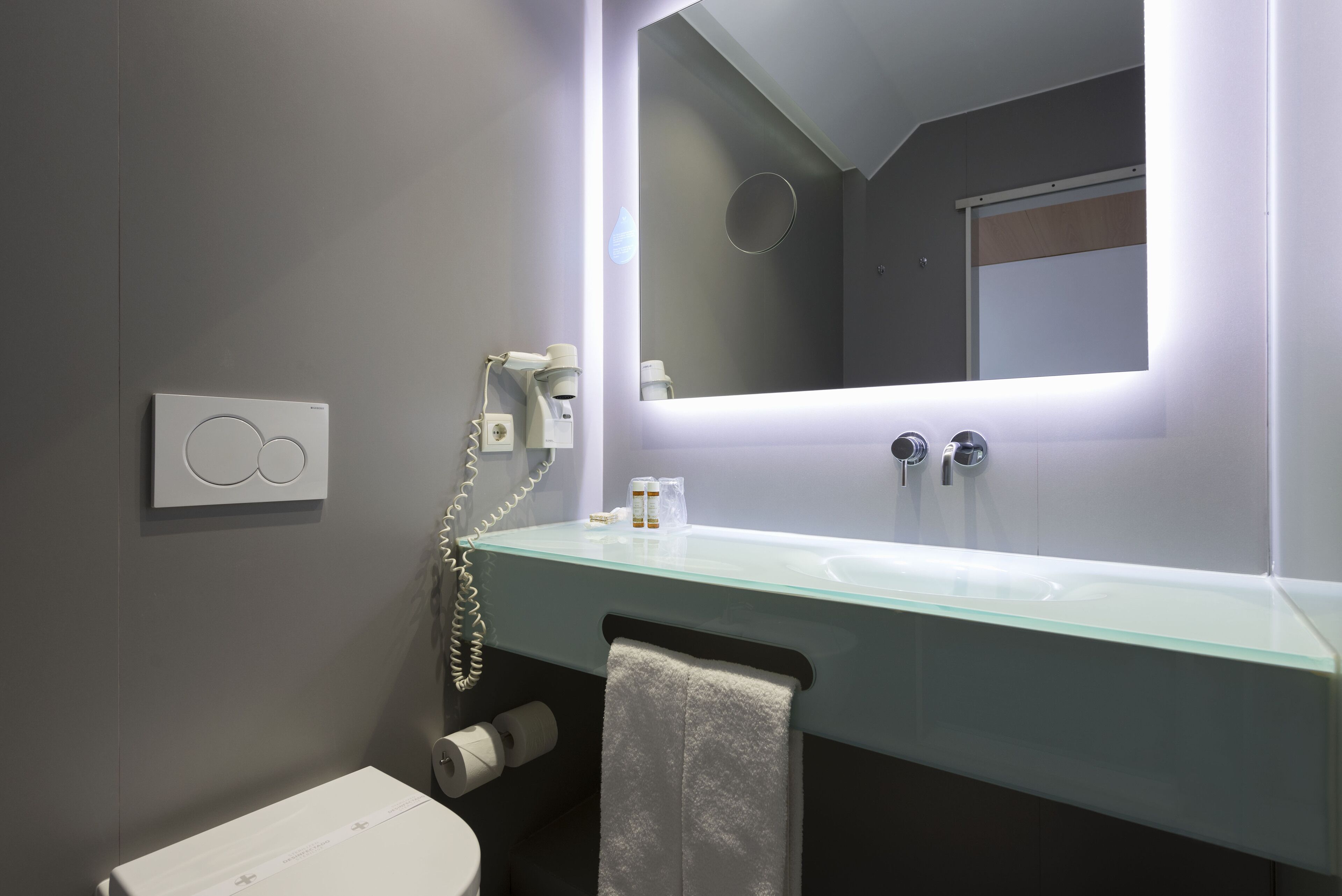 A modern bathroom with a glass sink, a towel hanging on the wall, a toilet, and a mirror with integrated lighting.