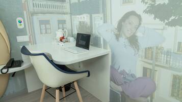 Ein Zimmer mit weißem Schreibtisch, einem Laptop, einem Stuhl, einem Telefon und einem Fenster mit Blick auf Gebäude.
