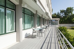 A covered outdoor seating area with tables and chairs, large windows, and a view of the surrounding greenery. - Varzinn Hotel (Povoa De Varzim)