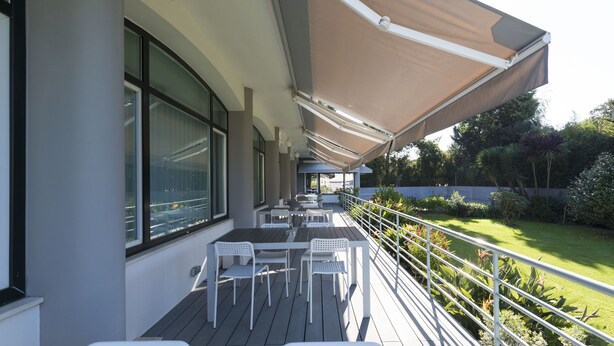 Une terrasse extérieure avec des tables et des chaises blanches, un grand auvent et une vue sur le jardin.