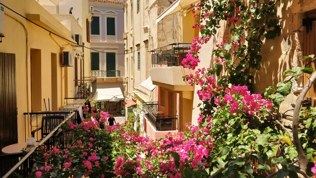 A narrow street with buildings on either side, balconies with railings, and a small outdoor seating area with tables and chairs. - Iason Studios (Chania)