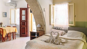 A bedroom with a bed, a dining table, and a chair. There is a window with lace curtains and a wooden door. - Iason Studios (Chania)