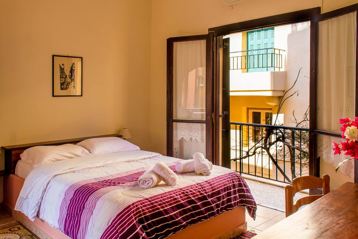 A hotel room with a bed, a chair, a table, and a balcony with a bicycle.