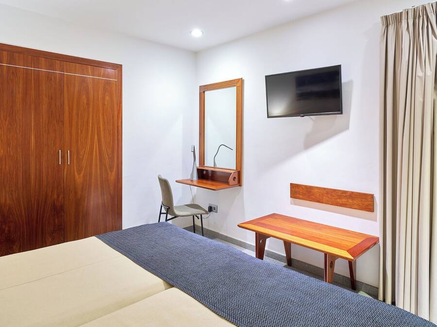A bedroom with a bed, a wooden wardrobe, a desk with a chair, a television, and a window with curtains.