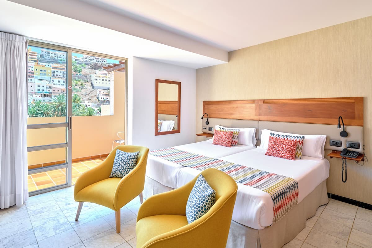 Una habitación de hotel con una cama grande, dos sillones amarillos, un balcón con vista a la ciudad y un teléfono en la mesita de noche.